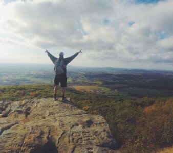 Man with backpack reaching arms to the clouds looking out over a landscape