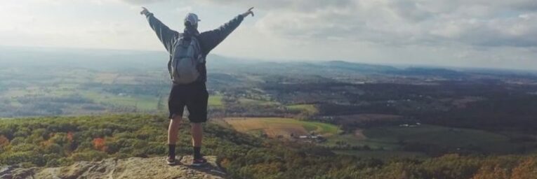 Man with backpack reaching arms to the clouds looking out over a landscape