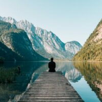man sitting on boardwalk staring at the mountains and water around him
